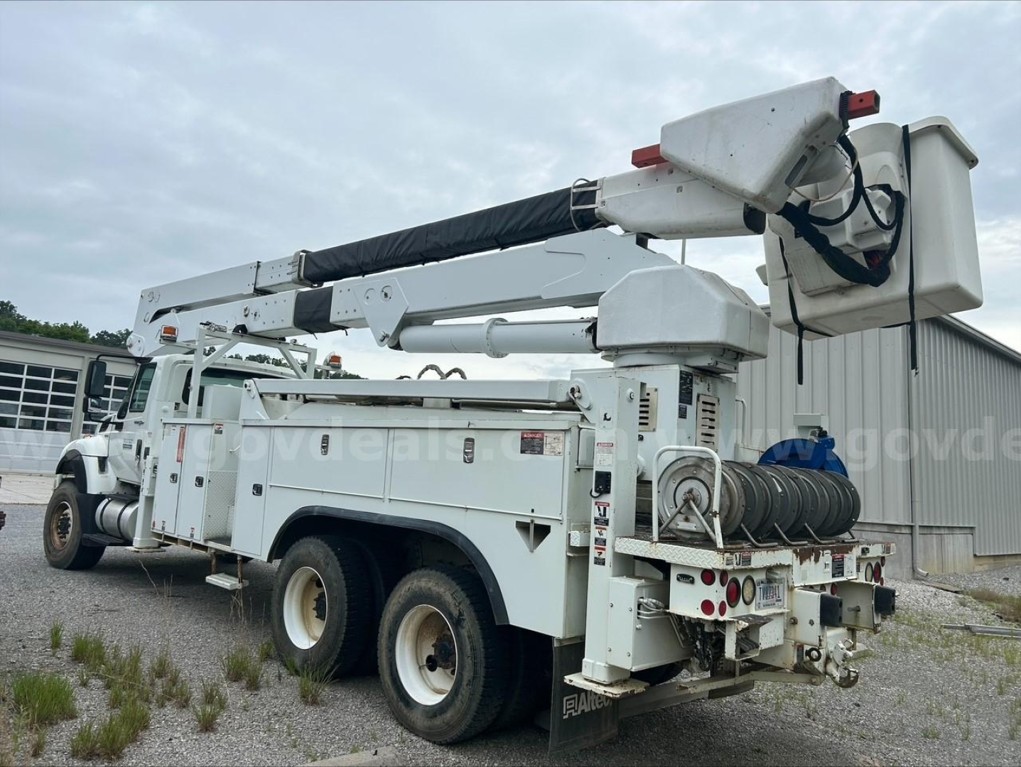 Platform bucket truck with white service body, boom lift extended and bucket at end. Various compartments and hose reels on sides; dual rear wheels; visible labels and decals.