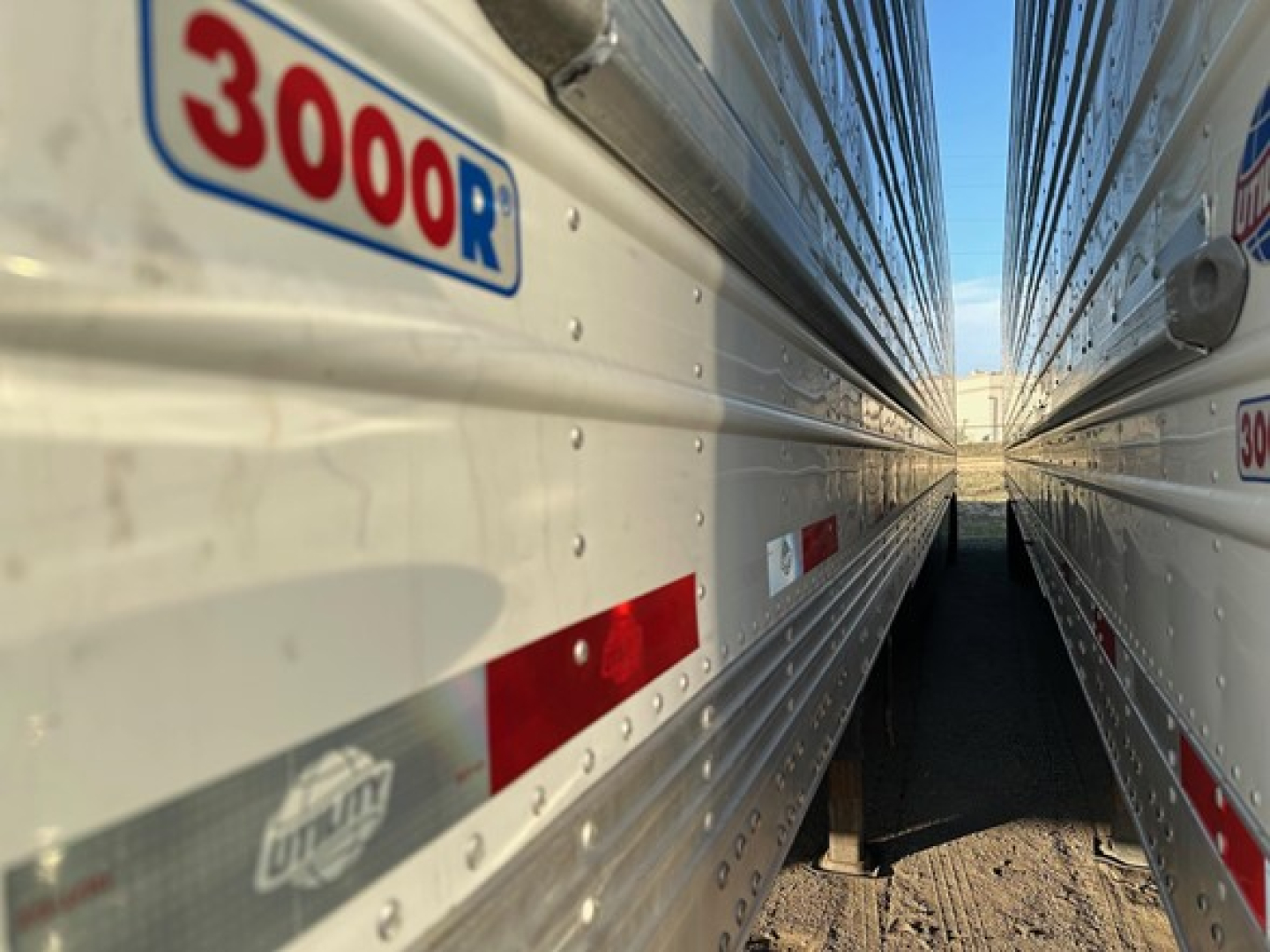 Sheeted refrigeration trailer side with '3000R' label and aluminum riveted wall, viewed between two trailers; partial UTMan logo and reflective red/white strips visible.