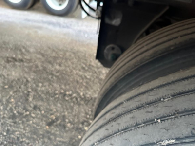 Trailer wheel and tire close-up view showing rubber tread and sidewall, with a portion of the trailer undercarriage and ground surface.