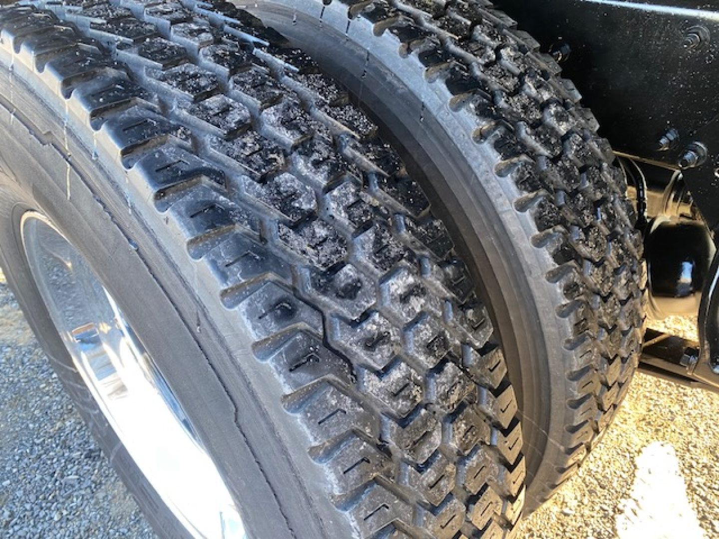 Freightliner M2-106 wheel and tire close-up showing dual rear tires with tread pattern and wheel/rim; part of flatbed truck undercarriage visible.