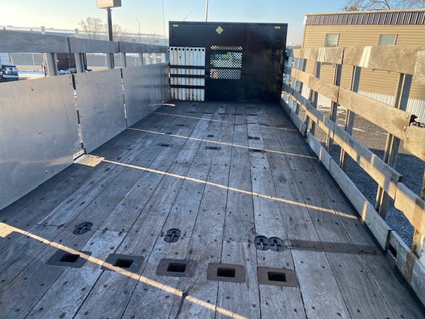 Flatbed truck bed of a Freightliner M2-106 with wooden planks, side stakes, and a black metal bulkhead at the cab end. VIN 3ALACXFC5LDLU3392 not visible on image but part of listing.