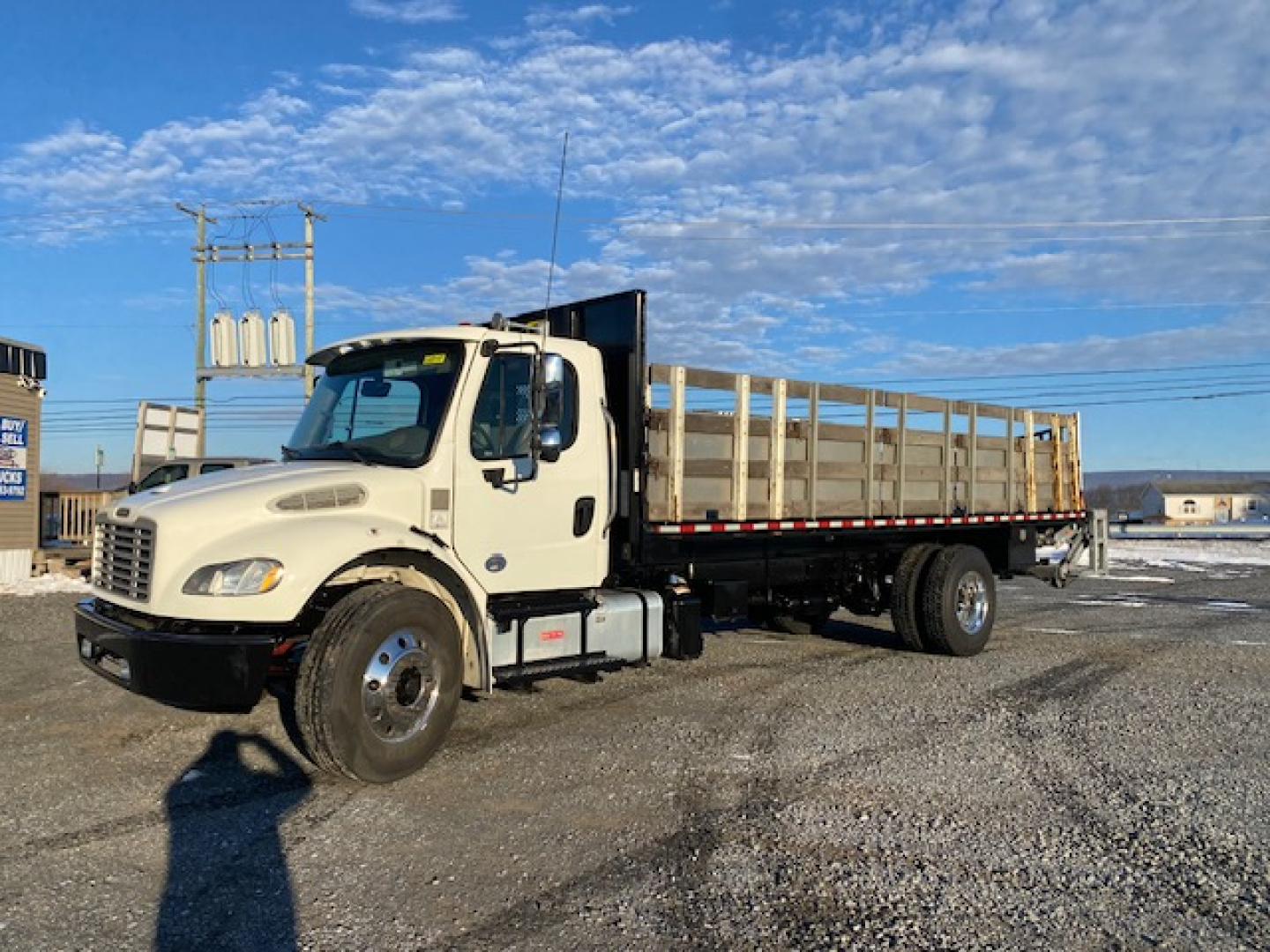 Freightliner M2-106 flatbed truck in a parking lot, side view showing cab and long dirt color flatbed with wooden stakes.