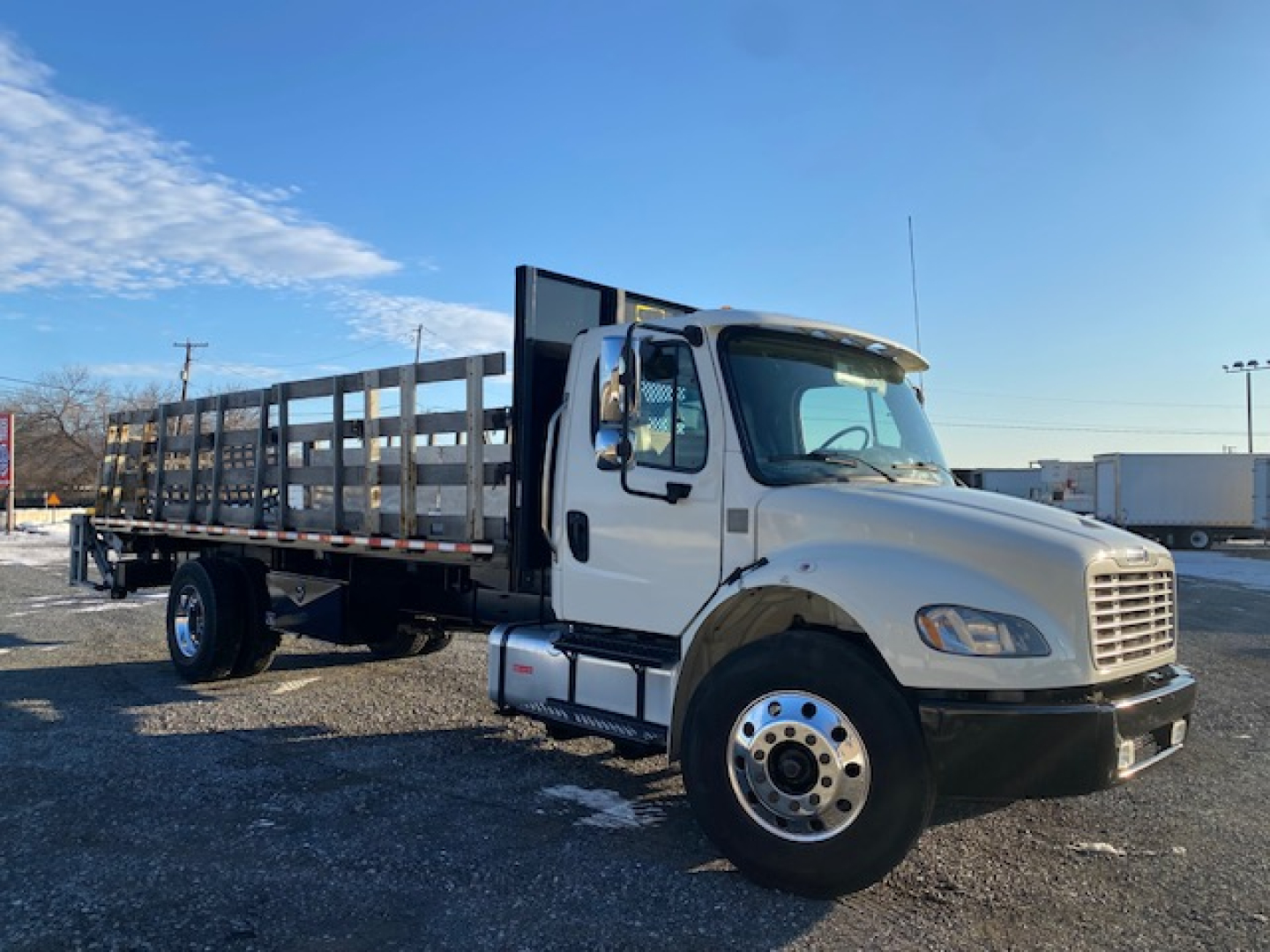 Flatbed truck showing Freightliner M2-106 cab with long steel stake side rails and open flatbed, front grille visible, white cab, daytime outdoor lot.