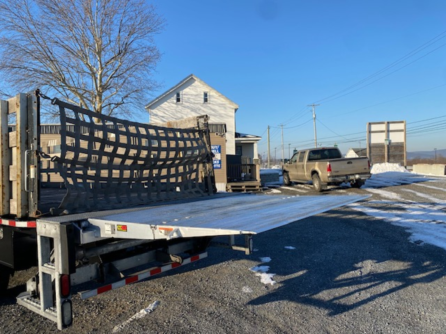 Flatbed tow truck with a sliced black mesh ramp on a trailer, a beige pickup in background, clear blue sky, snow patches on ground.