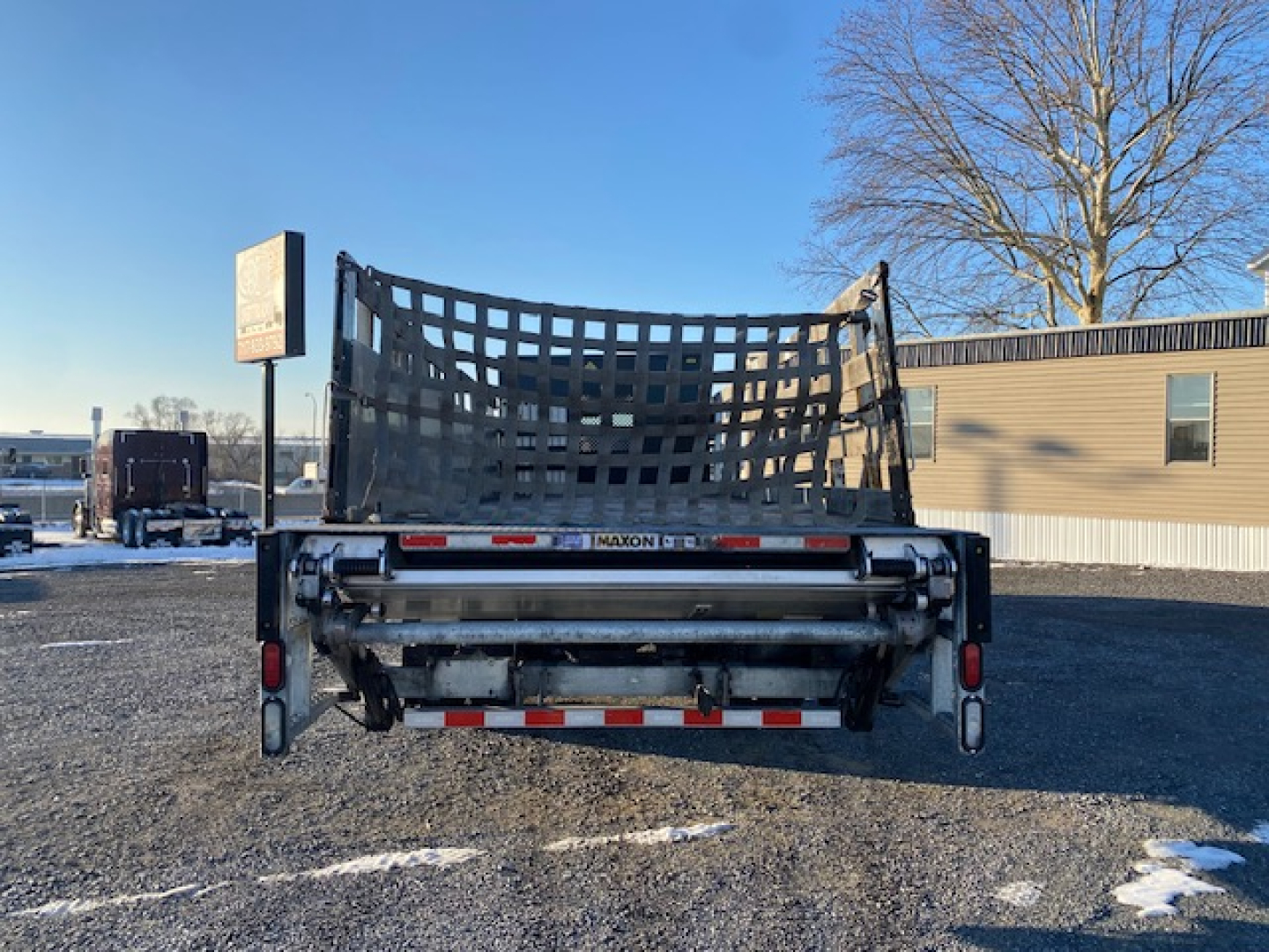 Freightliner M2-106 dump bed rear view showing black mesh tailgate and Maxon hydraulic mechanism, with reflective tape and lights.