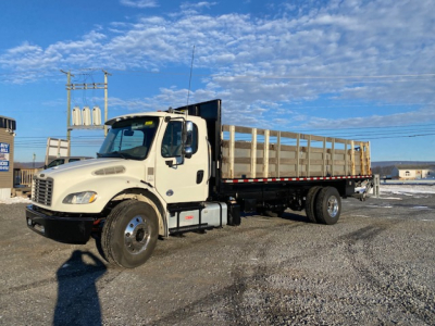 Freightliner M2-106 flatbed truck in a parking lot, side view showing cab and long dirt color flatbed with wooden stakes.