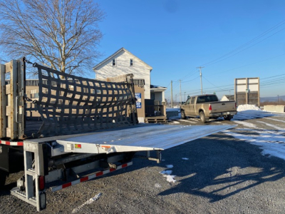 Flatbed tow truck with a sliced black mesh ramp on a trailer, a beige pickup in background, clear blue sky, snow patches on ground.