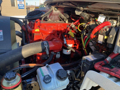 Freightliner M2-106 engine bay showing red diesel engine with hoses, coolant reservoir, and electrical wiring; various components visible under hood.