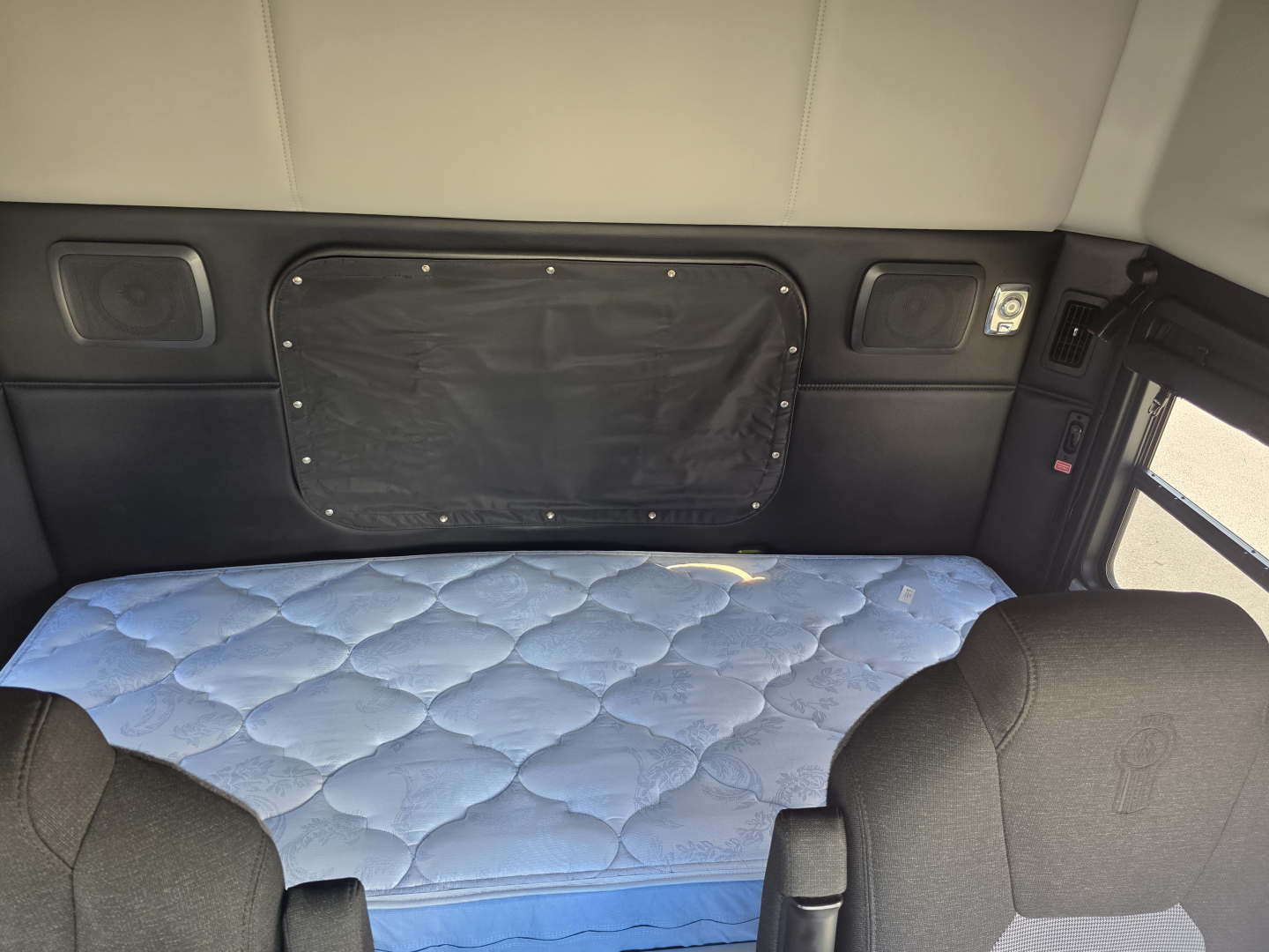 Kenworth T880 sleeper cab interior with a blue quilted mattress, black side panels, and overhead cabinet pockets visible in the sleeper area.