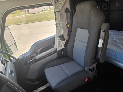 Truck cab interior showing driver seat and passenger seat with light curtains, dashboard and door panel; sleeper area bed visible in background.