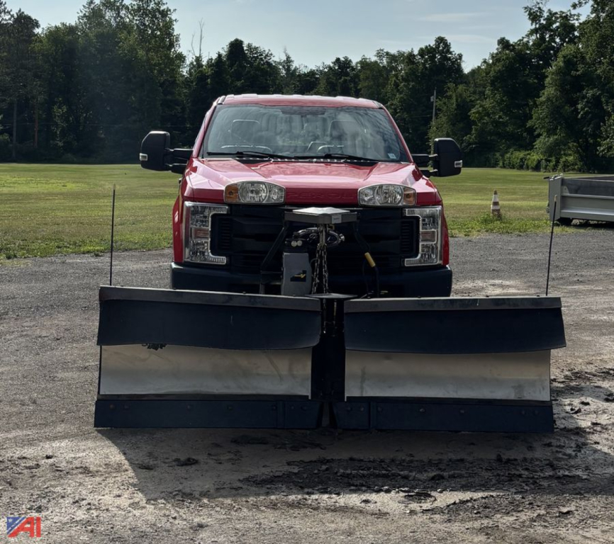 Front view of a red 2017 Ford F-250 Super Duty pickup with a large snow plow attachment mounted to the front, headlights visible, open area around plow, outdoor lot with grassy field in background.