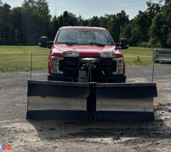 Front view of a red 2017 Ford F-250 Super Duty pickup with a large snow plow attachment mounted to the front, headlights visible, open area around plow, outdoor lot with grassy field in background.