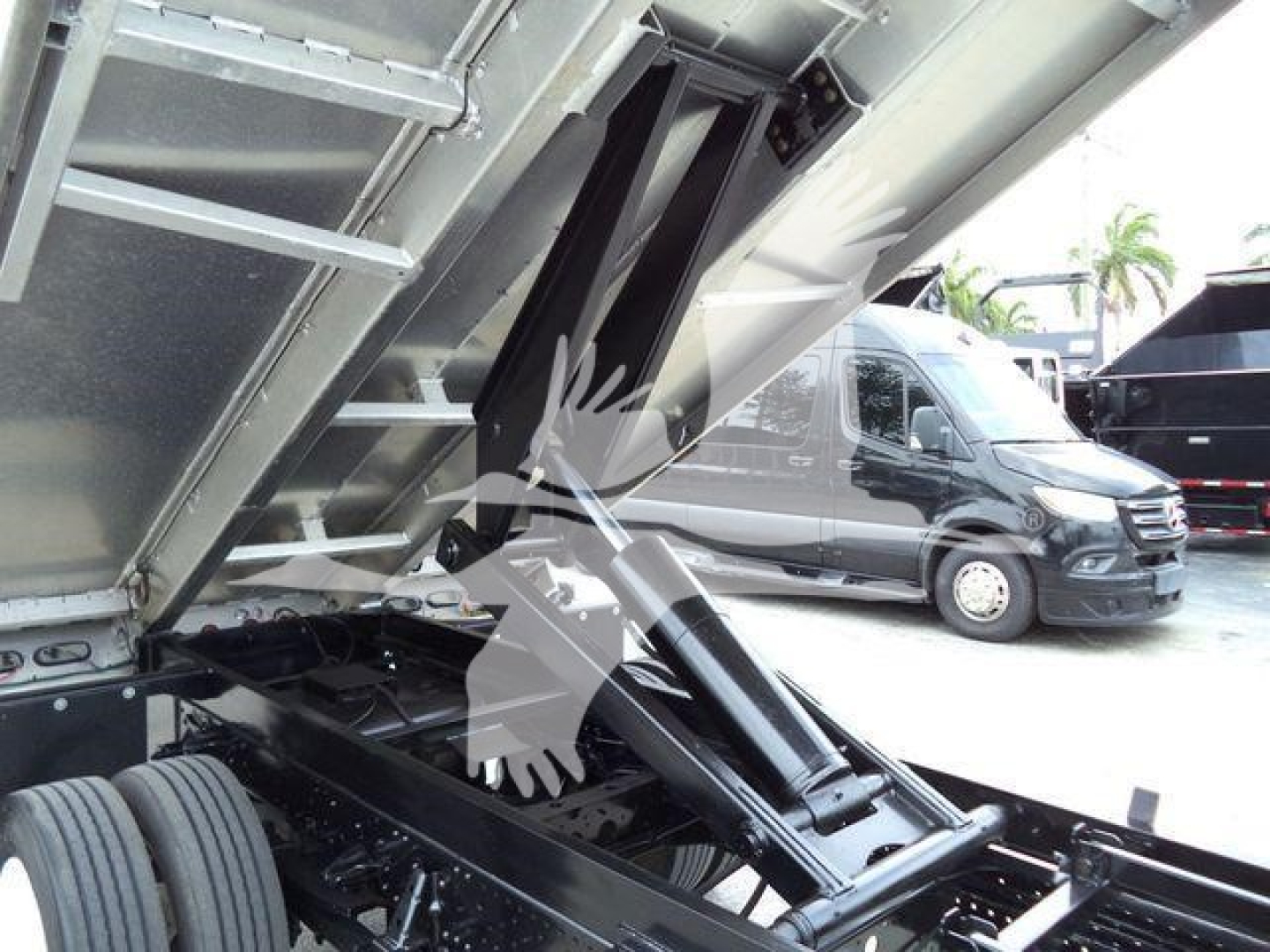Isuzu NPR HD truck bed interior showing hydraulic lift with rack and hinges, dark metal components, and a partial view of a side door and other trucks in a lot.
