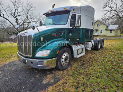 Peterbilt 579 semi truck in green and silver, front-left view, parked on grass with trees and a house in background.