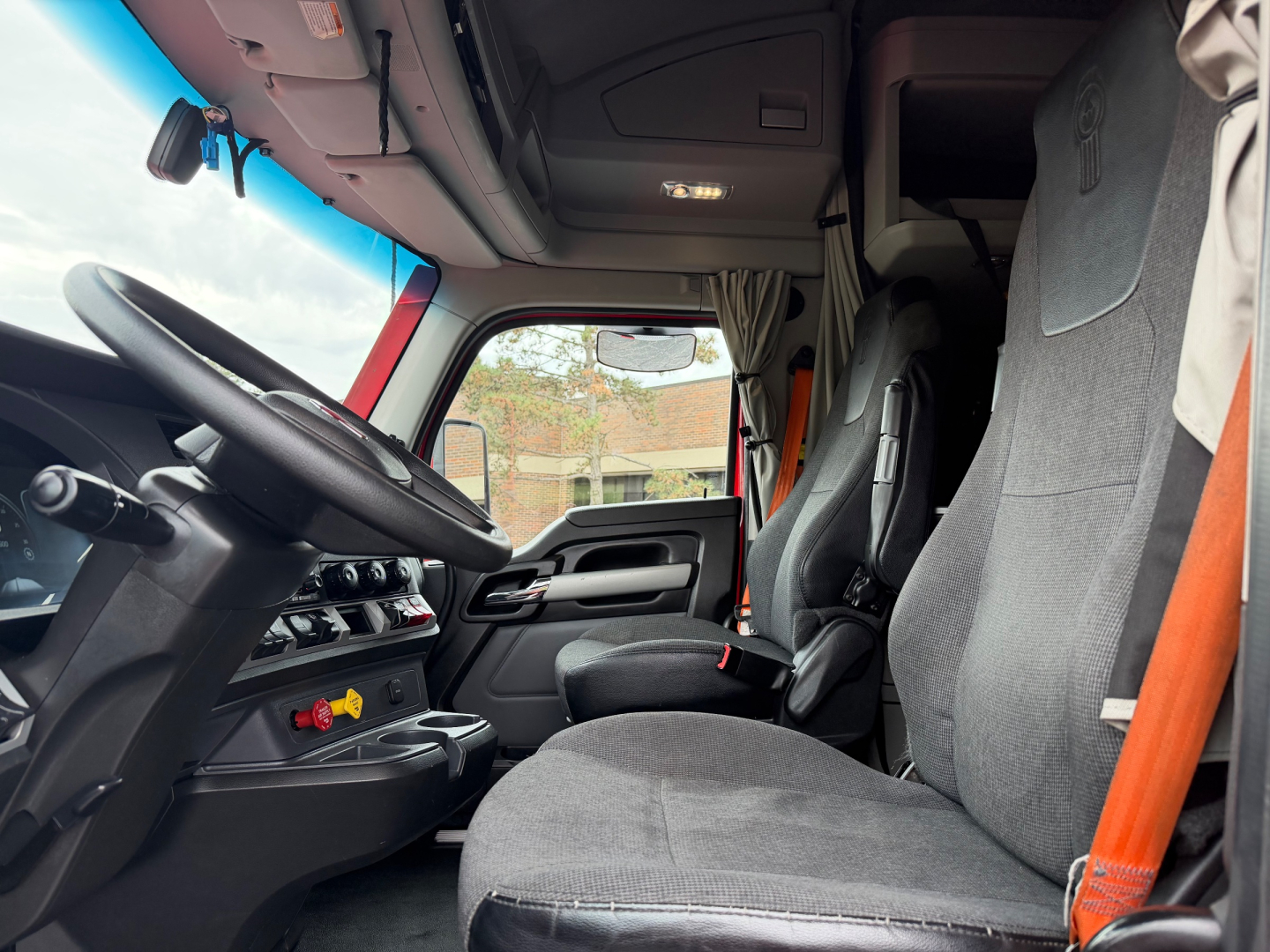 Cab interior of a Kenworth truck showing steering wheel, dashboard with controls, driver seat and passenger seat, grey fabric seats, sun visors, and orange seat belt.