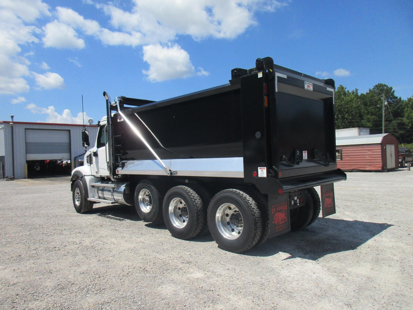 Dump truck rear and side showing Western Star 49X with three rear axles and a black dump body. Visible branding includes a small logo on the cab, chrome wheels, and a tailgate area.