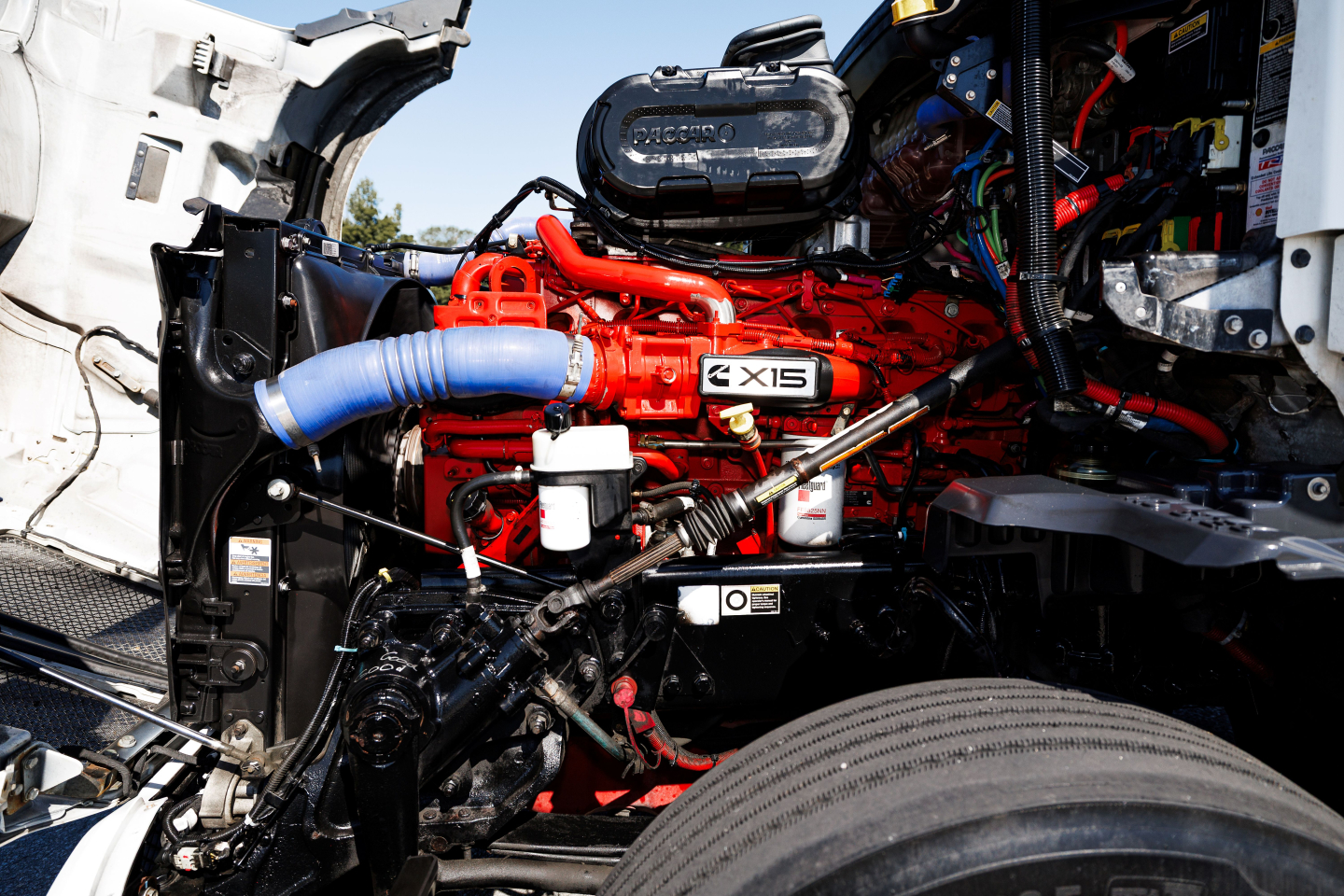 Kenworth T680 engine bay with bright red inline engine and blue hose visible, label GX15 on valve cover, various hoses and components.