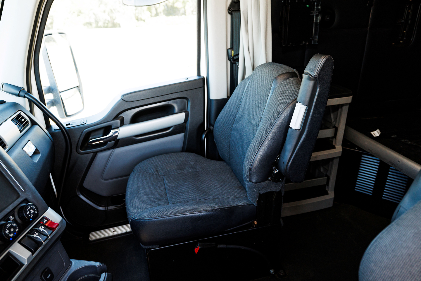 Truck cab interior: driver seat and passenger seat in view; dashboard area with steering wheel and door panel; part of sleeper area and storage shelves in background.