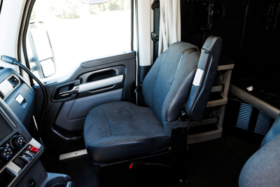 Truck cab interior: driver seat and passenger seat in view; dashboard area with steering wheel and door panel; part of sleeper area and storage shelves in background.