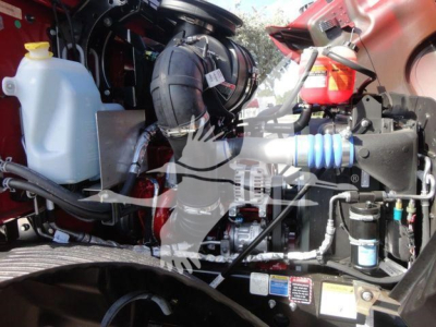 Engine bay of a new Mack MD6 rollback truck showing coolant reservoir, air intake hose, blue air line, and various engine components.