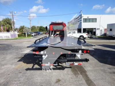 Tow truck flatbed with rollback configuration, rear view showing metal deck and hitch, red cab in distance and clear sky.