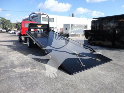 Flatbed tow truck with a lowered loading ramp and red cab, in a dealership lot, showing a DOT-style flatbed and support equipment.