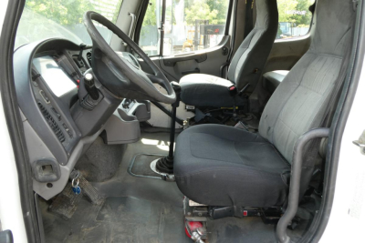 Truck cab interior of a Freightliner M2 112 showing steering wheel, driver seat, and dashboard with gear shifter and pedals.