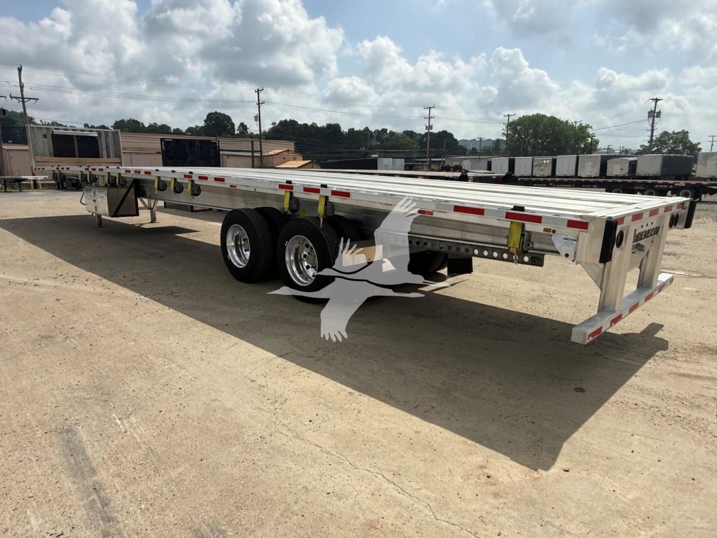Aluminum Benson flatbed trailer showing dual rear axles and flatbed frame with side rails and rear bumper, parked on gravel.