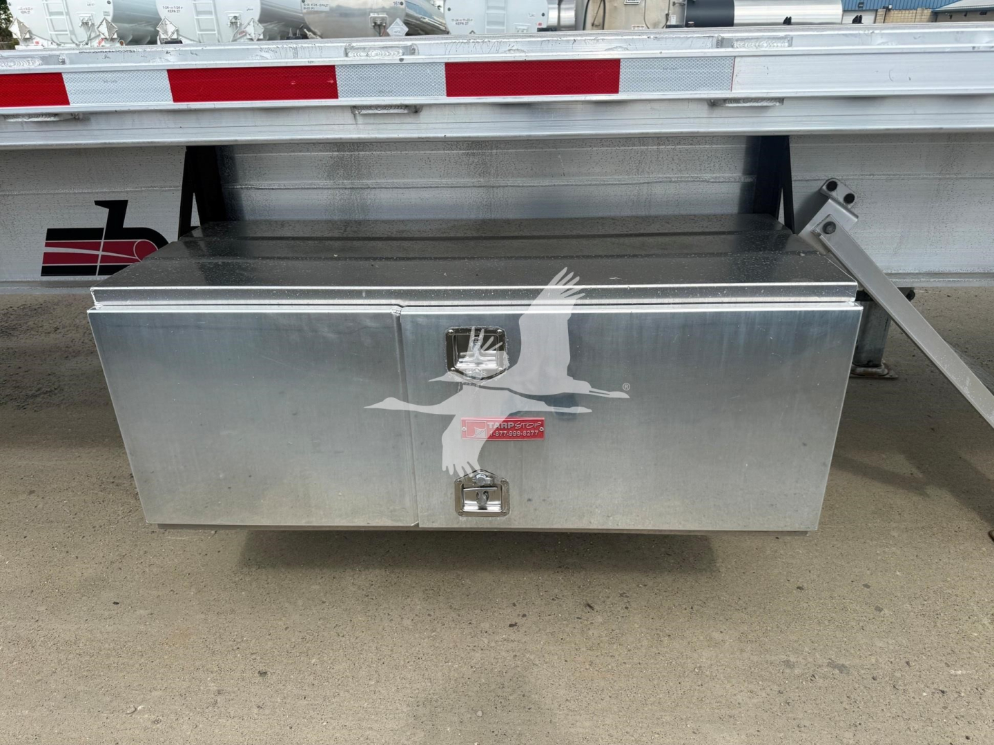 Flatbed trailer toolbox mounted under trailer with aluminum diamond plate lid and two latching handles.