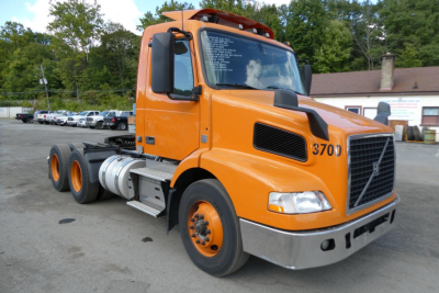 Orange Volvo VNM64T conventional daycab truck with 3700 badge on hood, large front grille, black side mirrors, and dual rear axles; text on windshield lists specs.
