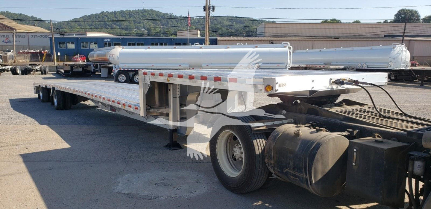 Flatbed trailer in a lot showing a long deck and tandem axle set with cylindrical fuel tanks and winch components visible.