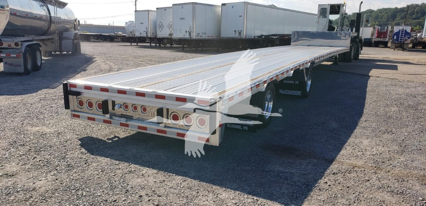 Flatbed trailer with long aluminum deck and rear lights, parked in a lot; rear bumper with circular brake lights and white reflective trim.