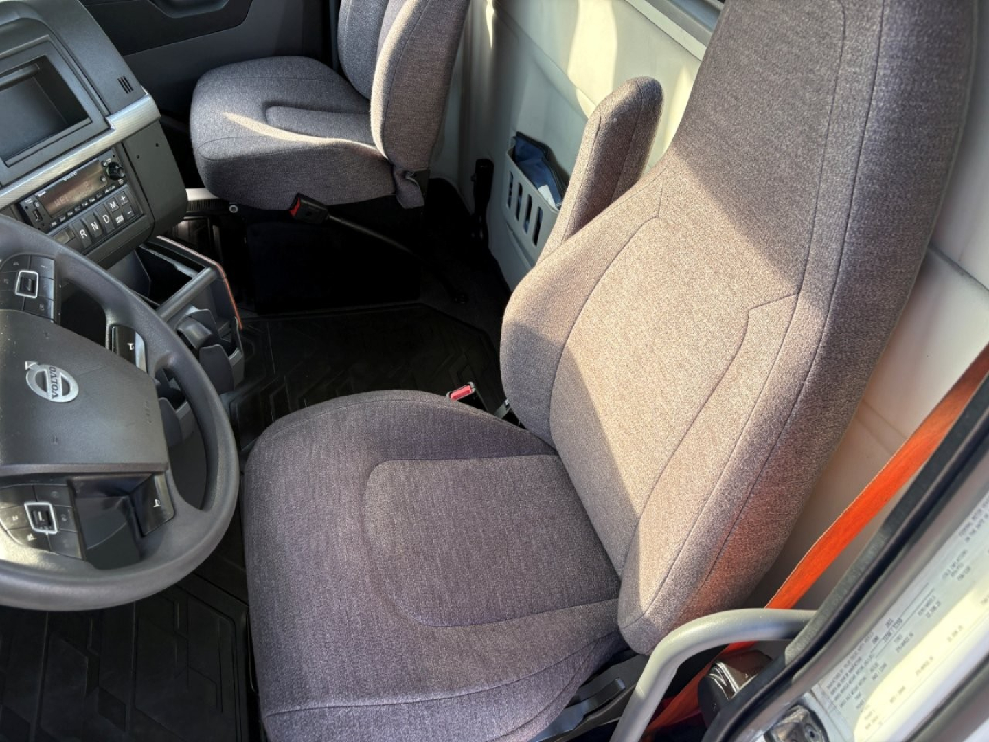Cab interior of a Volvo VNR truck showing driver seat, steering wheel with Volvo logo, and passenger seat; grey fabric seats with center console and dashboard visible.