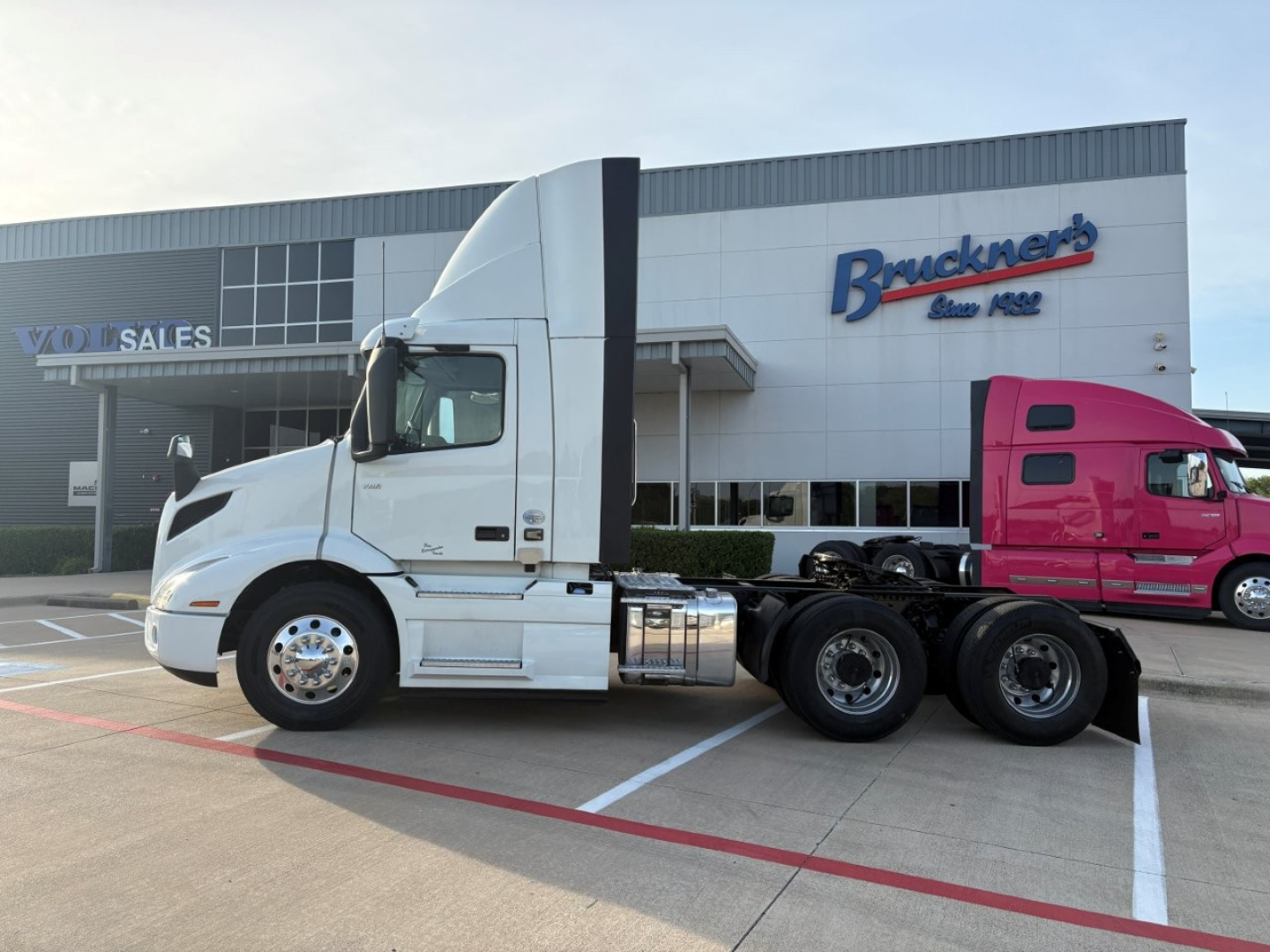 Truck in lot showing a white Volvo VNR64T300 daytime cab with 6x4 rear dual axles, parked in front of a dealership building with Bruckner’s sign.