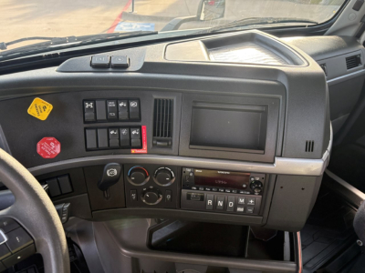 Dashboard of a Volvo truck cab showing controls and a screen; a yellow caution sticker and red hazard sticker are visible near switches.