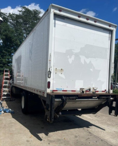 Box truck rear view showing white Hino 268 cargo box with closed roll-up door, bumper hitch, and undercarriage chassis.