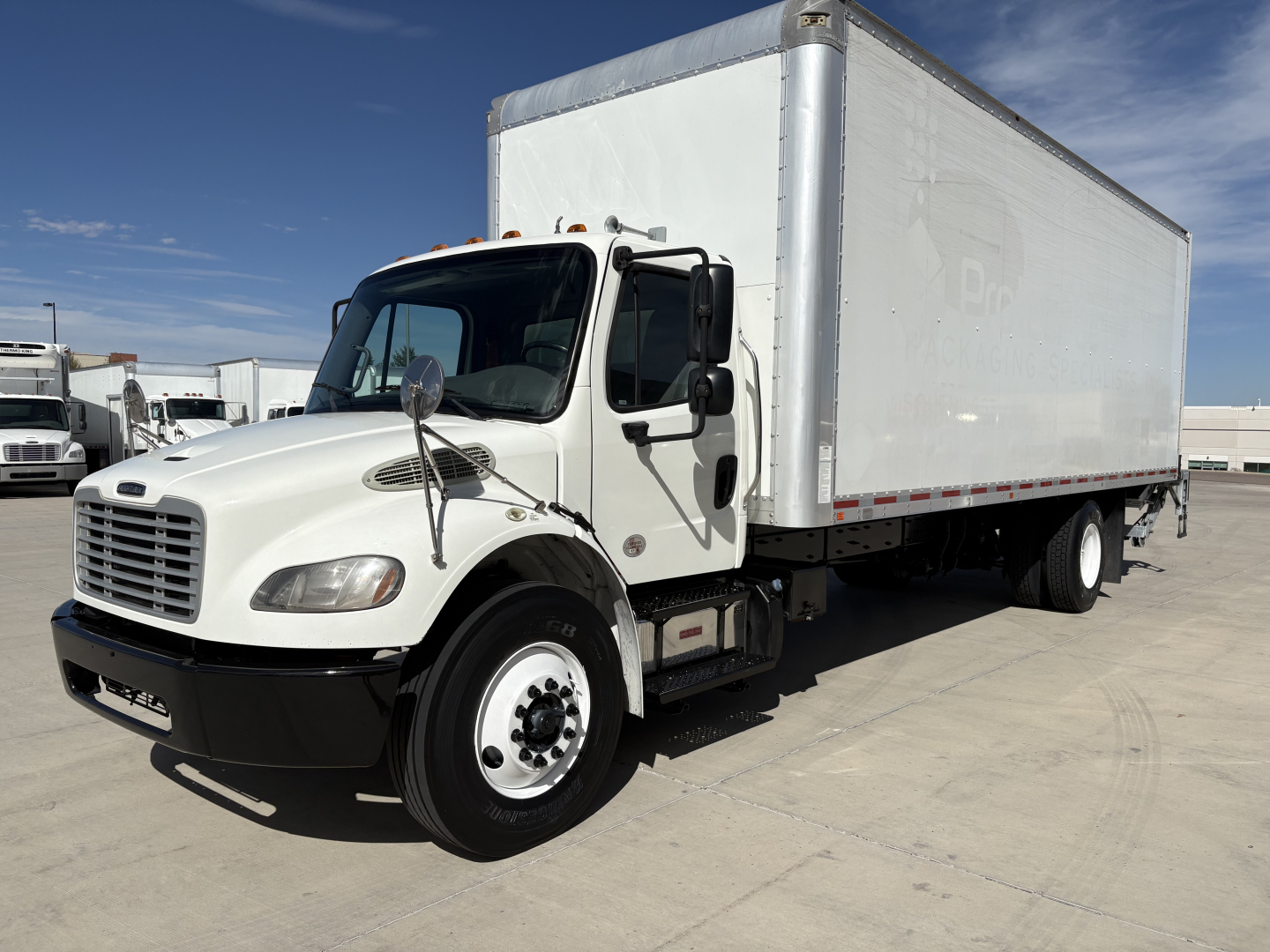 Box truck in a freightliner m2-106 with white cab and large box trailer, side view showing front and passenger side, clear sky.
