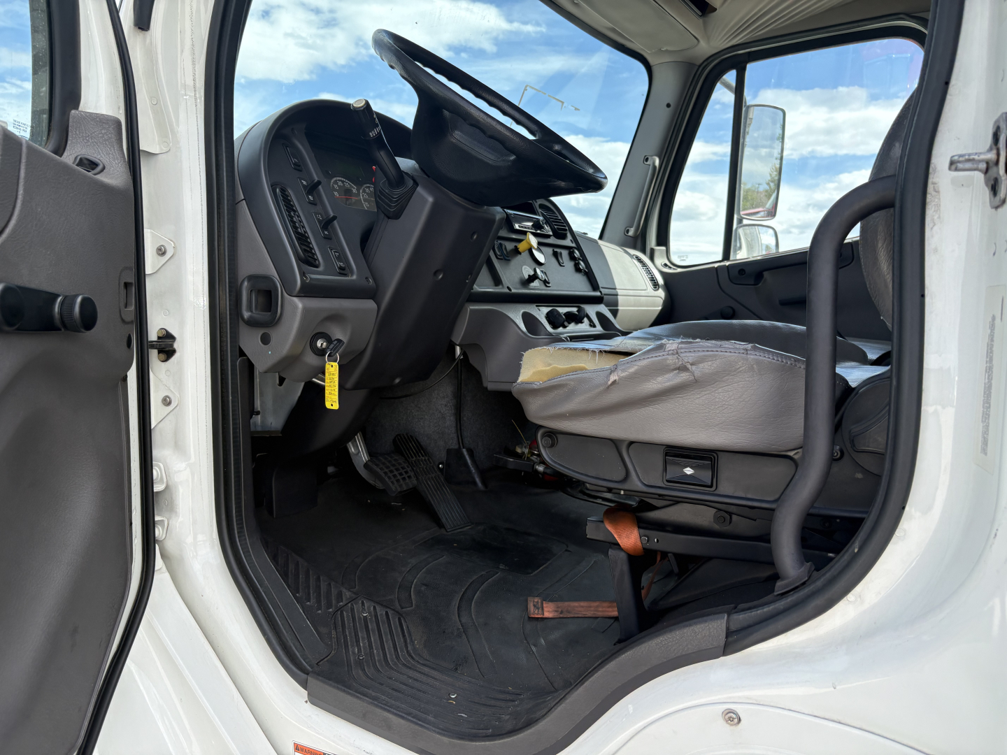 Cab interior of a Freightliner M2-106 with steering wheel, dashboard, and driver's seat visible from open door.