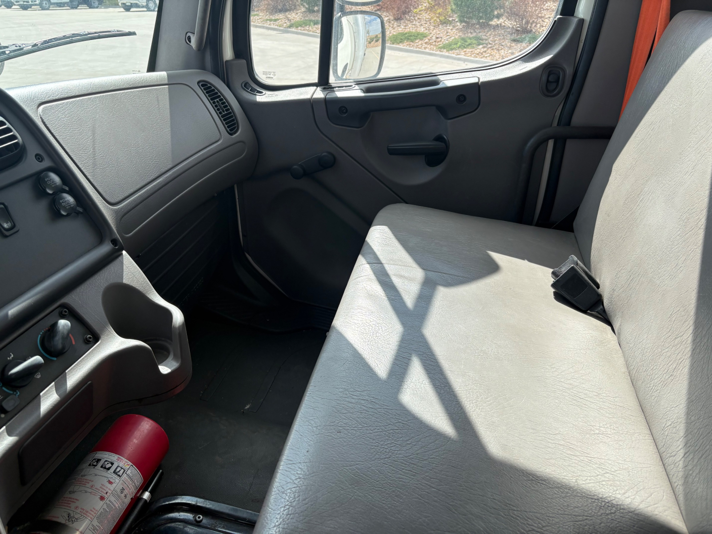 Cab interior of a Freightliner M2-106 truck with gray bench seat, steering panel, and window, showing a red fire extinguisher at the floor. Visible text includes vehicle details such as VIN, stock number, category, type, and condition from