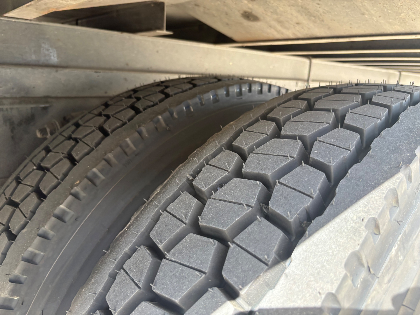 Freightliner M2-106 wheel and dual tires with tread pattern visible under truck frame; metal suspension components and frame rail above.