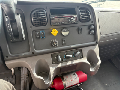 Freightliner M2-106 interior dashboard view showing HVAC controls, steering column, cupholders, and a red fire extinguisher under the dash.