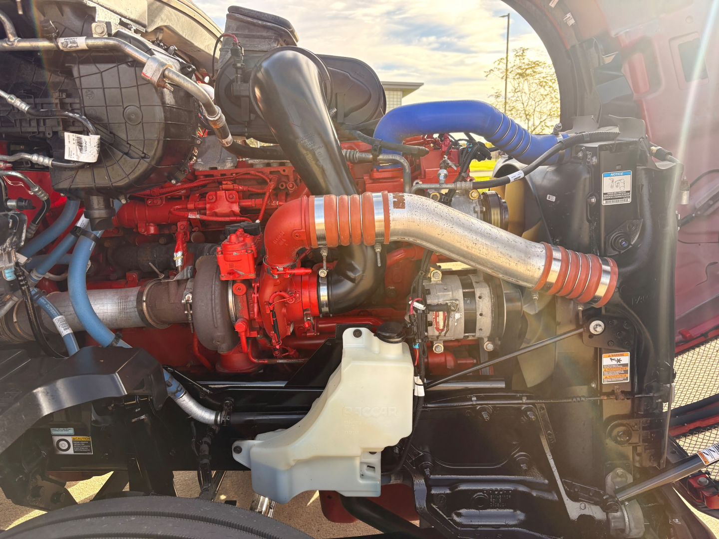 Kenworth T680 engine bay showing red engine block, large exhaust pipe, blue silicone hose, coolant reservoir, and various hoses and clamps.