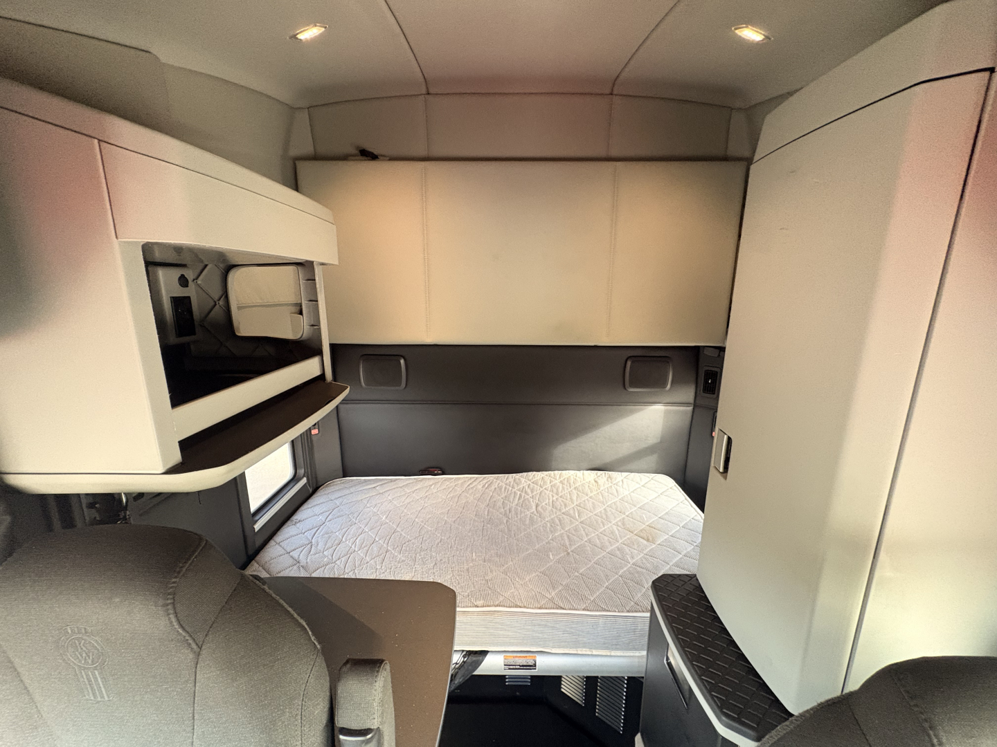 Interior cab view of a 2023 Kenworth T680 truck sleeper area with a raised bed, overhead storage, and compact control panel; clean and well-lit cabin.