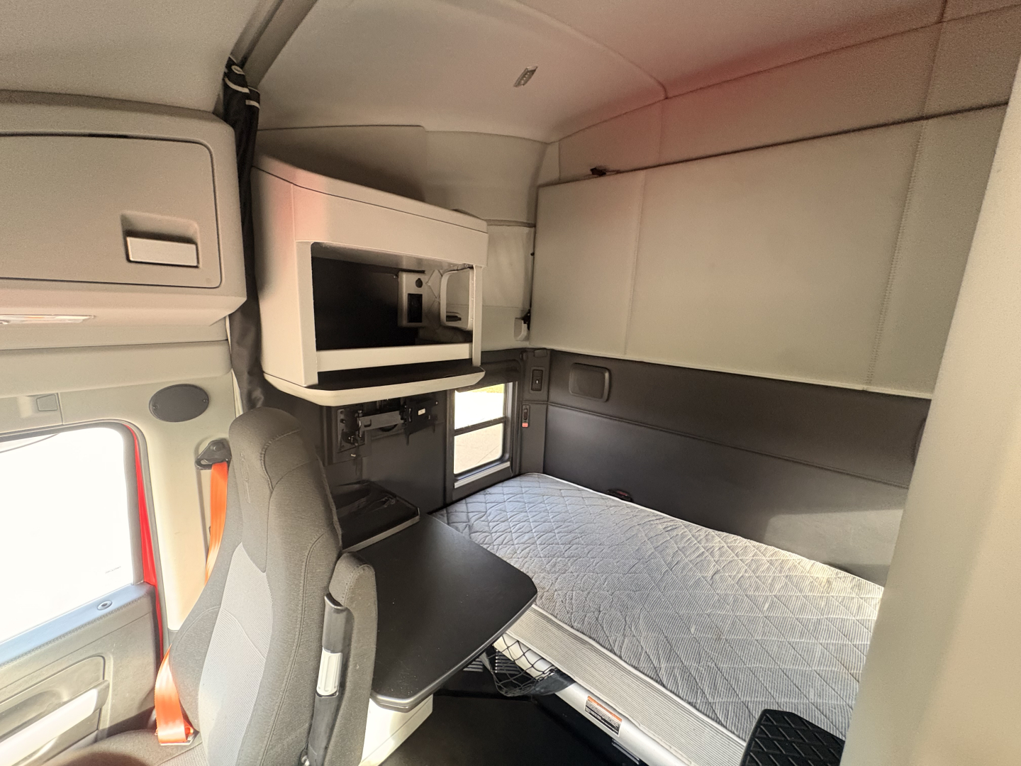 Kenworth T680 sleeper cab interior with bed and workspace, cabin storage above, and window visible.