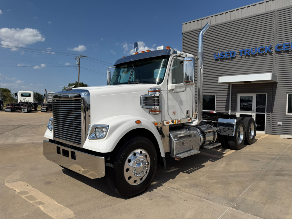 Freightliner heavy truck cab and chassis in a dealership lot, chrome mirrors, silver fuel tanks, and a large vertical grille. Visible text on the building reads USED TRUCK CENTER.