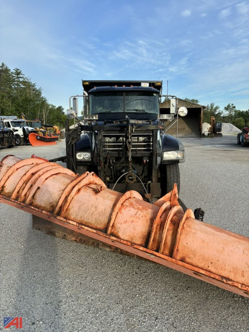 Dump truck with plow attachment facing camera; Mack Granite 64FR dump truck in service with orange conveyor/plow blade in foreground; truck cab and equipment visible.