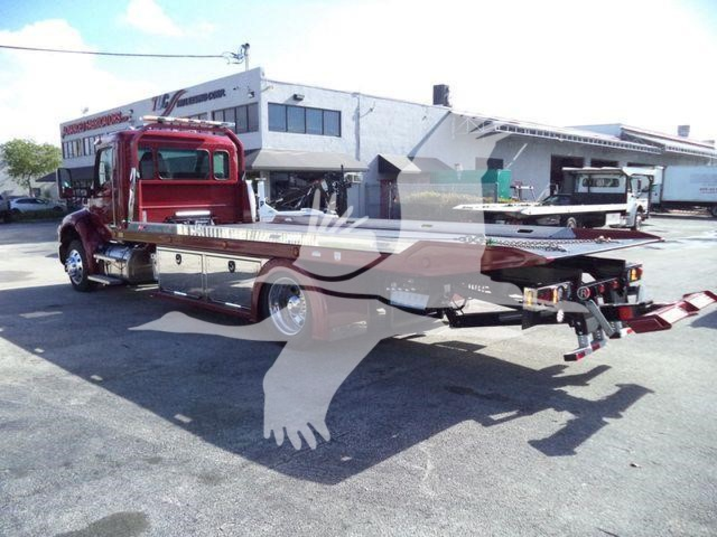Kenworth T280 rollback tow truck with extended flatbed and wheel lift, red cab, dealership lot in background, visible chrome exhaust and hydraulic components.