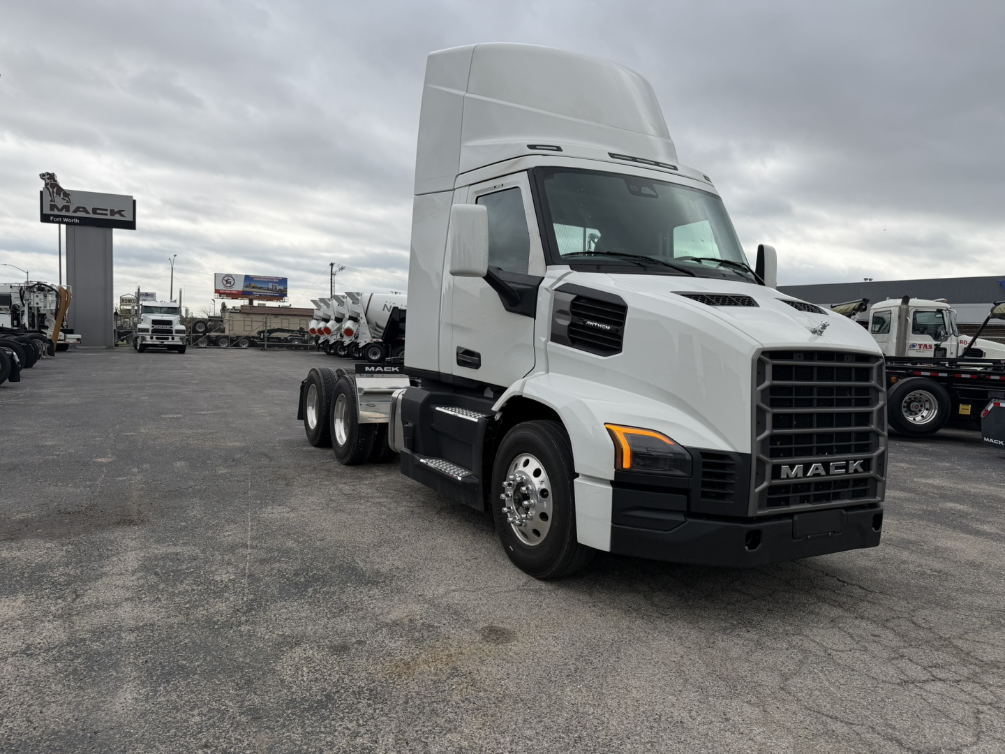 Mack AN64T conventional daycab truck in a dealership lot, white exterior, high sleeper, front grille with Mack logo, multiple trucks in background.