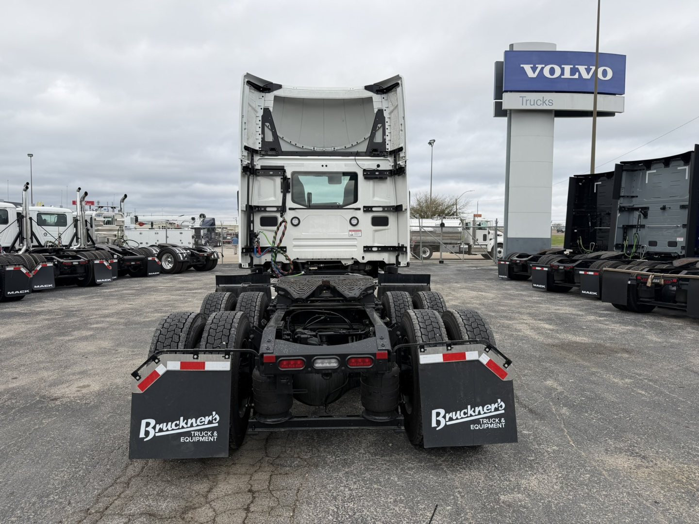 Truck chassis rear view with fifth wheel, mud flaps labeled 'Bruckner's Equipment' and a Mack AN64T cab visible in distance. Volvo Trucks sign in background.