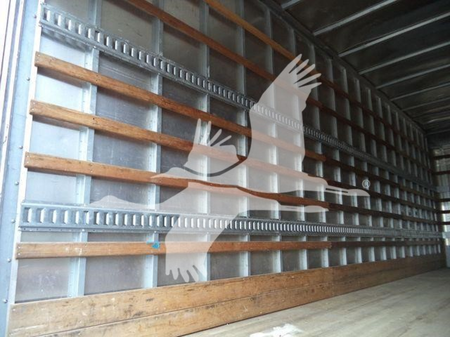 Inside view of a Freightliner M2 106 bare trailer interior with metal sidewalls and wooden rails showing no cargo; watermark of a stylized truck logo obscures part of the scene.